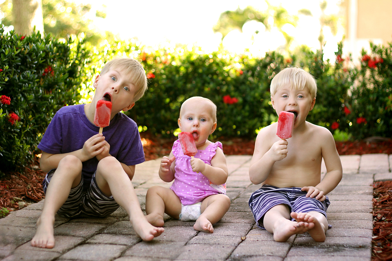 The Truth About Sugar: How It Affects Your Teeth at Every Age, Three young children, including two brothers and a baby sister are sitting outside in the garden, eating strawberry fruit popsicles on a summer day.